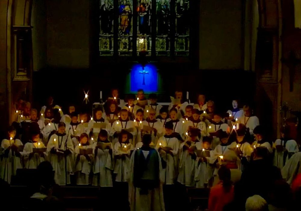 Robed choir stands in darkness, holding candles, in front of the high altar in church, facing the congregation, with director of music conducting. 
