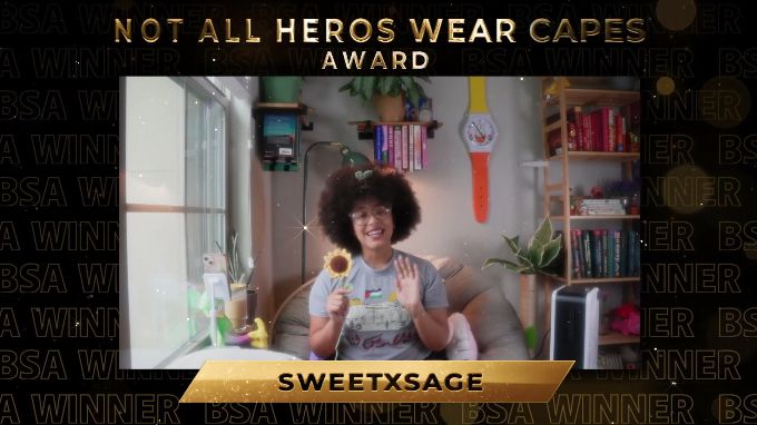 sage is sitting in a large round chair in a corner surrounded by books, plants and a large window. Sage is holding a crochet sunflower and is wearing a sprout in their hair. The image of sage in their stream room is edited on top of a black background that reads 'winner' over and over in gold. At the top of the image are the words 'Not All Heros Wear Capes Award'. at the bottom reads 'sweetxsage' in gold.