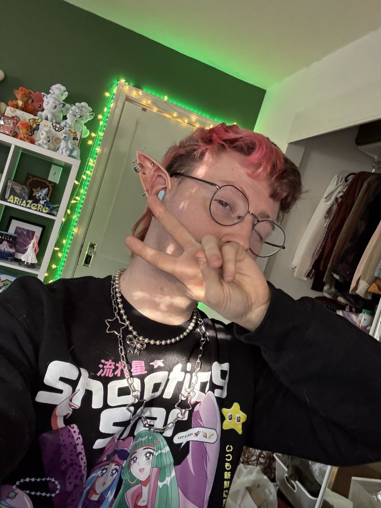 Aria Zero out of drag wearing a black sweatshirt with an anime graphic, silver star/bow necklaces, and elf ears! Their hair is half red, half brown and they are holding up a peace sign.