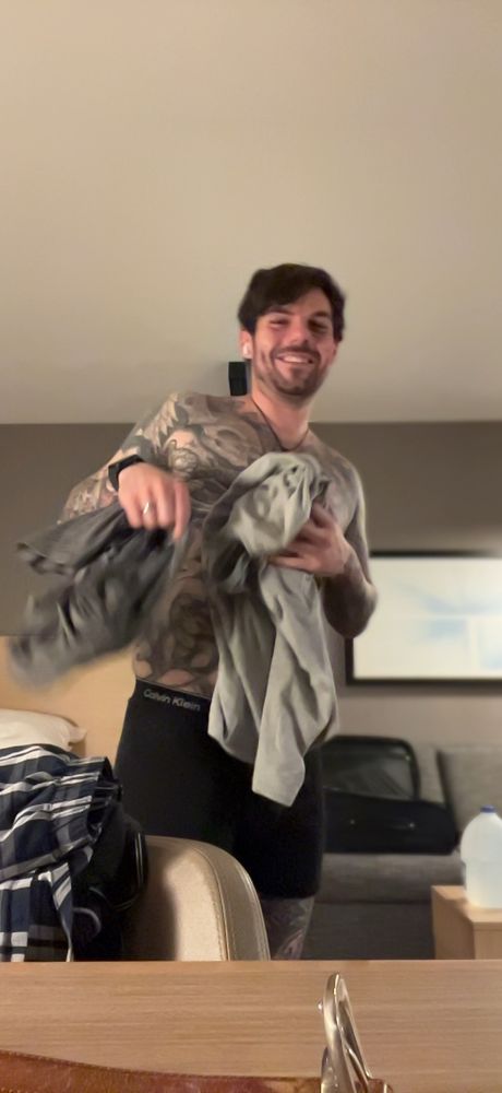 photo of a facetime photo taken of a tattooed male standing in a hotel room while undressing and smiling at the camera 
