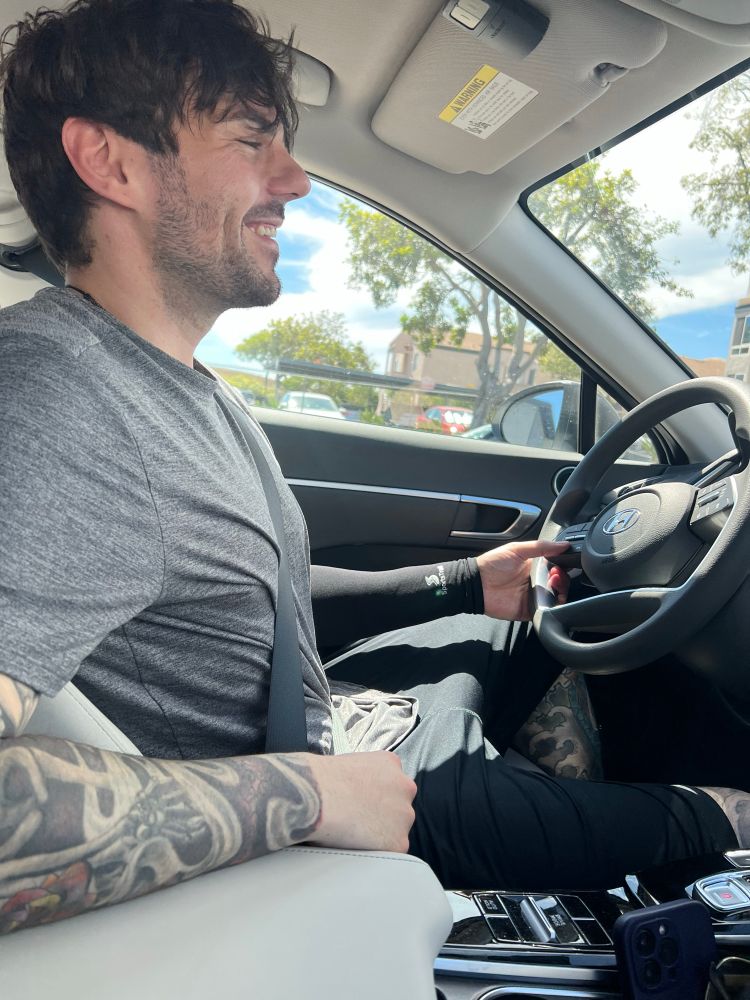 photo of a tattooed male sitting in the drivers seat of a car and smiling 