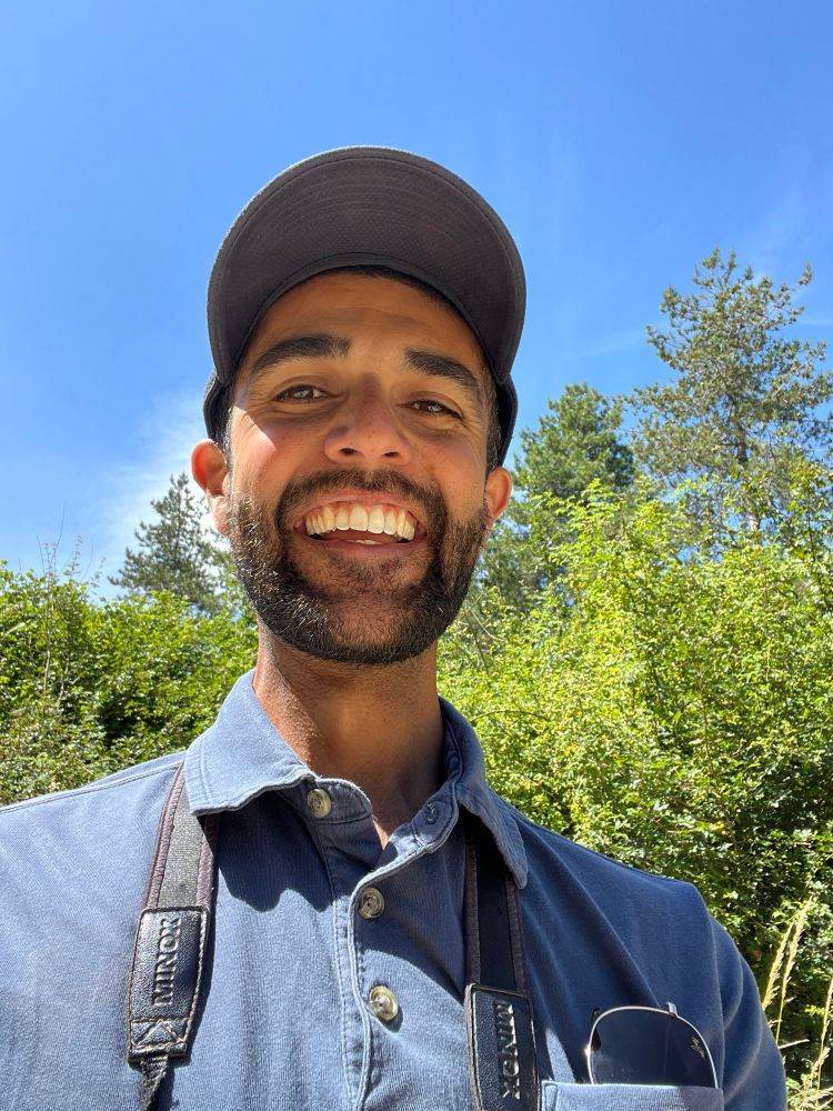 Selfie of man with big grin, stood in an area of woodland with blue sky above. He is wearing a baseball cap and has binoculars around his neck