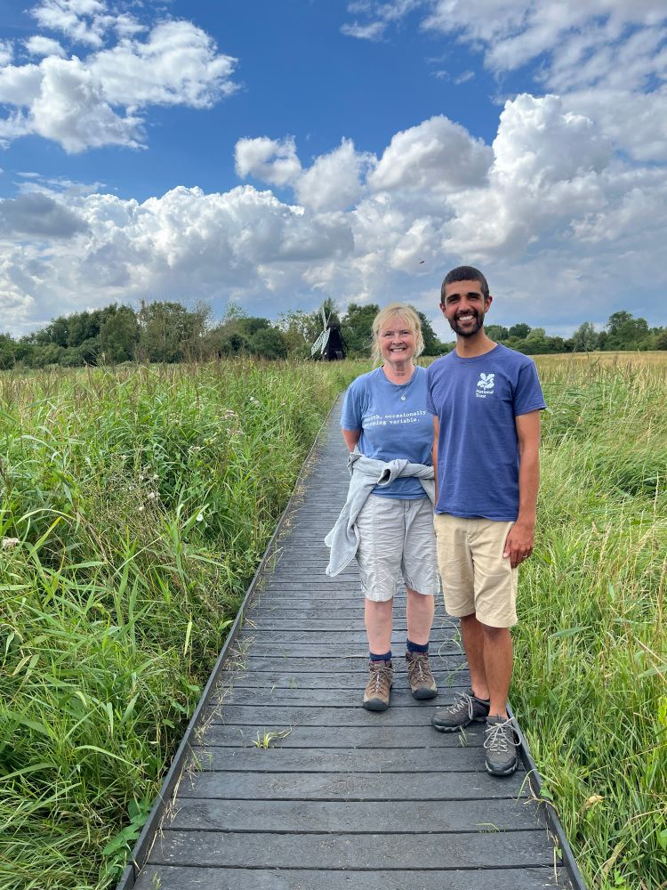 Ajay and Martha stood side by side on boardwalk both are wearing blue t-shirts and light shorts. A windmill is visible on the distance with lush green vegetation either side of the boardwalk, blue sky and white clouds above