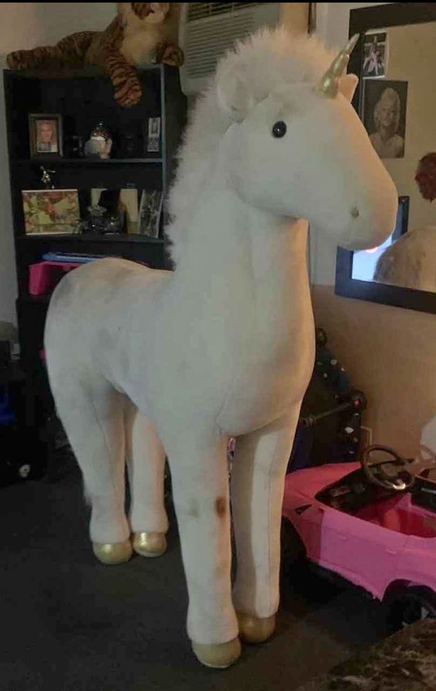 A very large unicorn with a rust colored stand on the knee. Behind the unicorn is a black bookshelf at least 6 feet tall. 