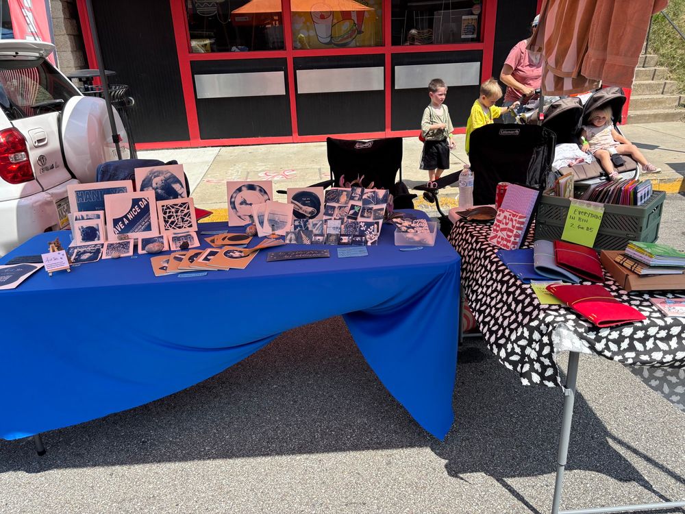 A booth at an art show showing one table of blue and white prints and magnets, carefully arranged and another of handwritten signs on like, notebooks and journals and a loose piece of ghost fabric 