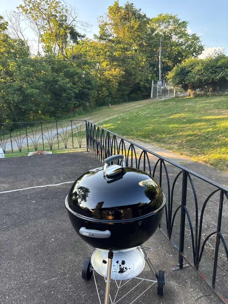 A large Weber charcoal grill on a patio and then behind that in the shade of the trees there is a full sized deer loafed.