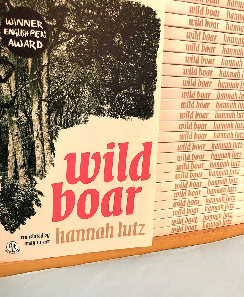 ID: Wild Boar cover against Wild Boar Spines. Cream cover with green, illustrated forest and red text. 