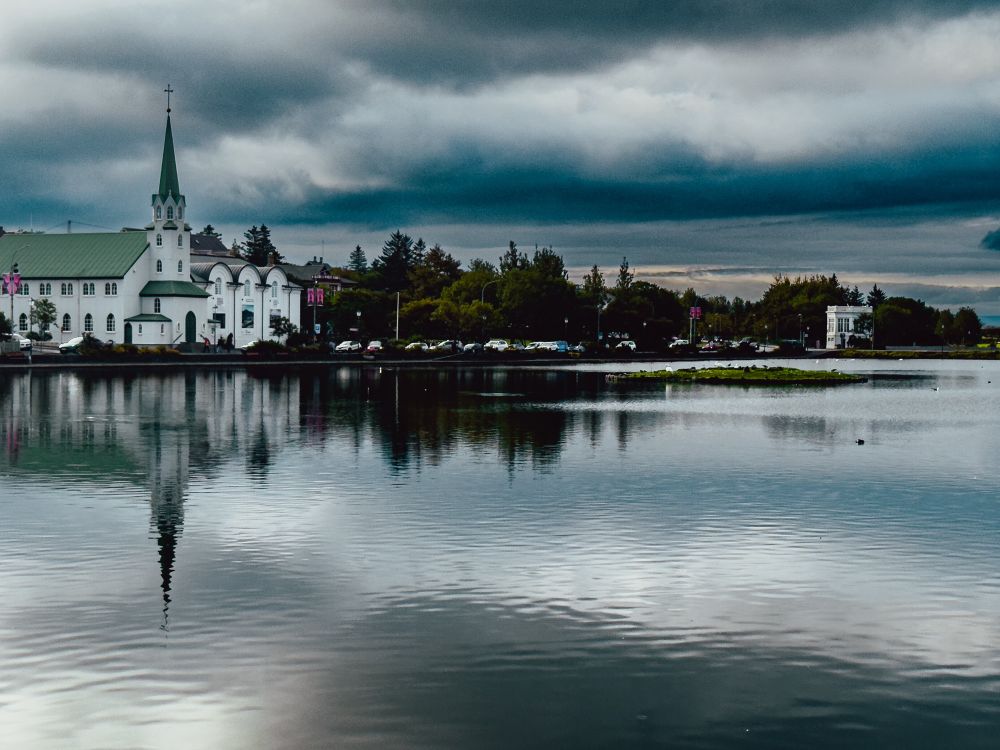 A white church reflected in the grey waters of a lake, beneath moody skies 