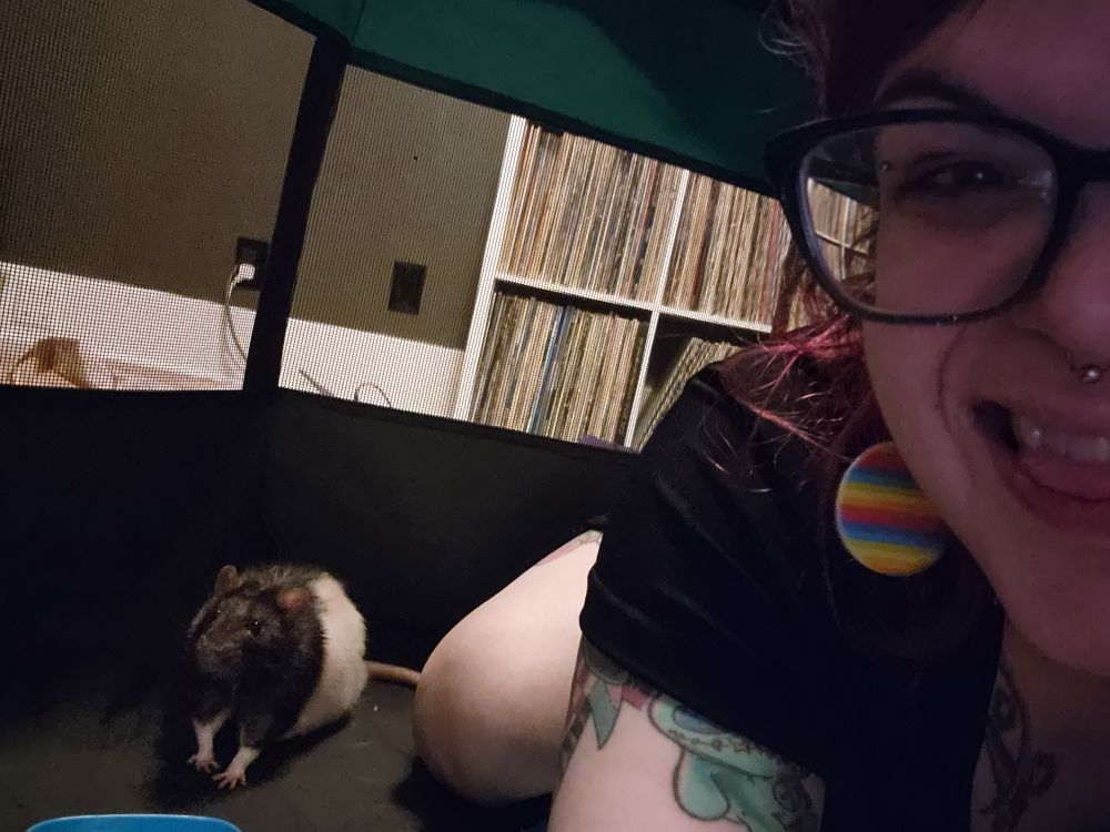 a smiling person (half out of frame) with glasses sitting beside a small brown and white rat