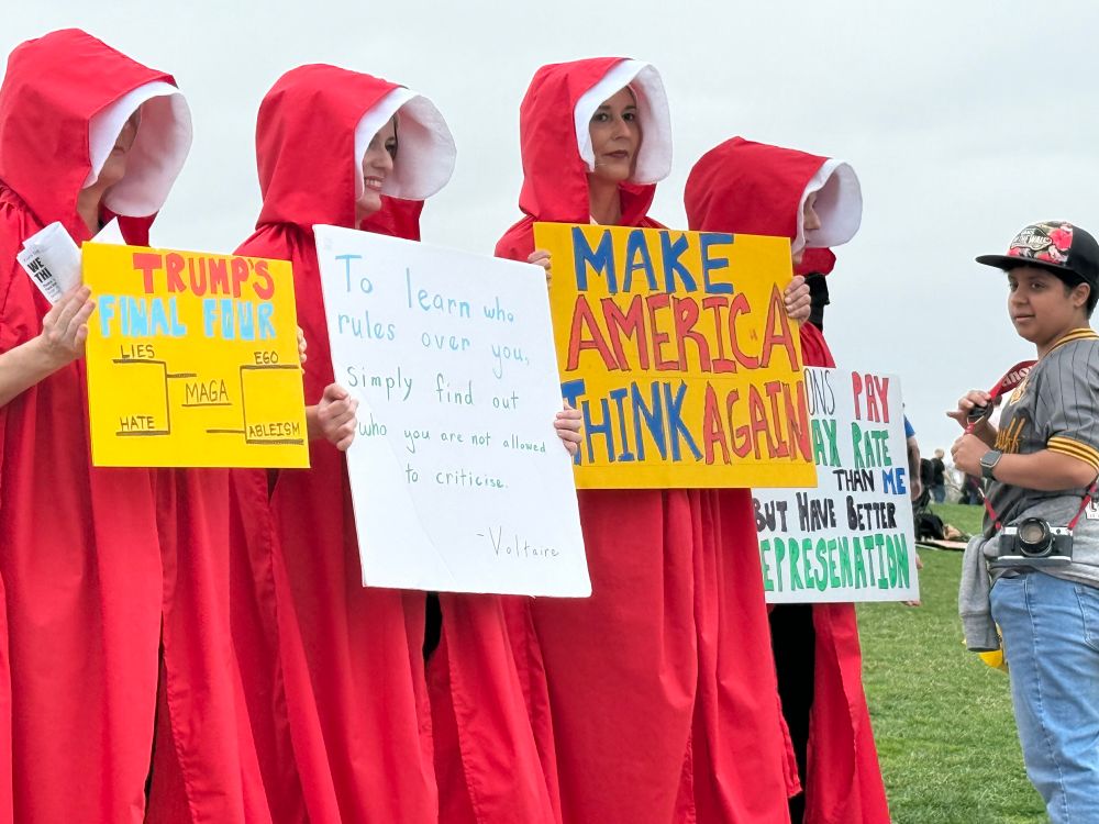 Women dressed from the Handmaid’s Tale with several signs. One says, “To learn who rules over you, simply find out who you are not allowed to criticize. Voltaire” 