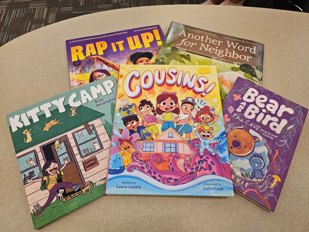 Five picture books spread on a table from top left: Rap It Up! by Carole and Jeffrey Boston Weatherford with Ernel Martinez, Another Word for Neighbor by Angela Pham Krans with Thai My Phuong, Kitty Camp by Drew Brockington, Cousins! by Laura Lavoie with Luisa Leal, and Bear and Bird The Cave and Other Stories by Jarvis.