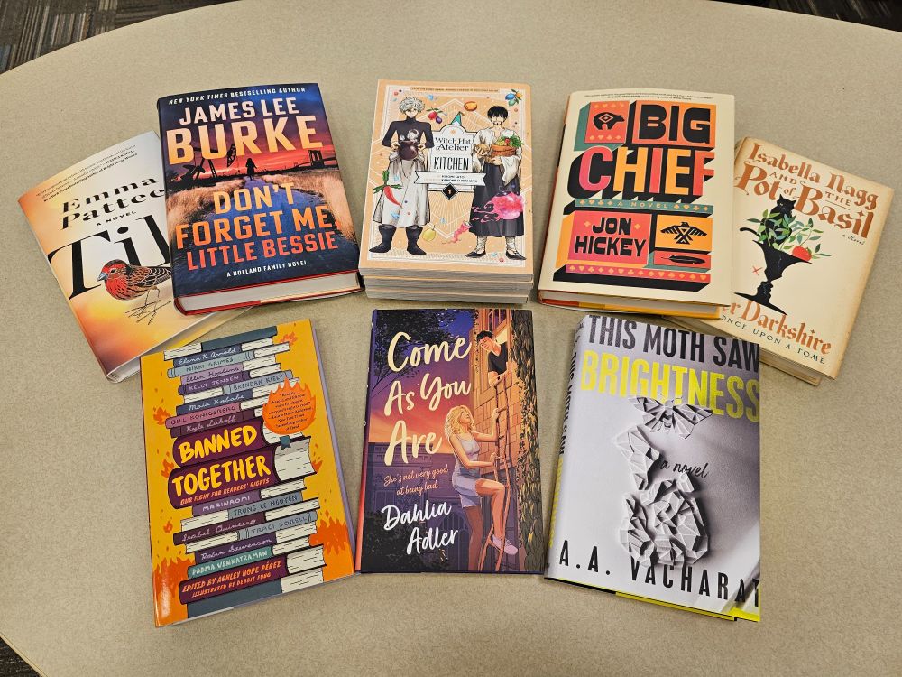 Eight titles spread on a table, from top left: Tilt by Emma Pattee, Don't Forget Me, Little Bessie by James Lee Burke, Witch Hat Atelier Kitchen volumes 1-5, Big Chief by Jon Hickey, Isabella Nagg and the Pot of Basil by Oliver Darkshire, Banned Together edited by Ashley Hope Perez, Come as You Are by Dahlia Adler, and This Moth Saw Brightness by A.A. Vacharat.
