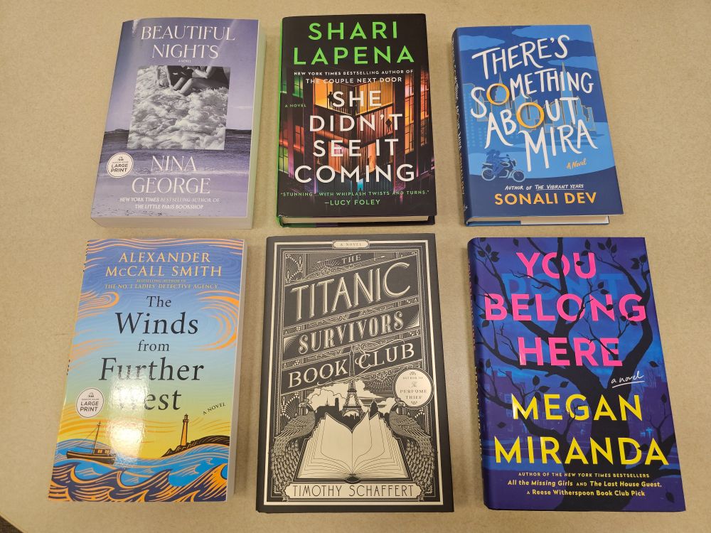 Six fiction books on a table, from top left: Beautiful Nights by Nina George in large print, She Didn't See It Coming by Shari Lapena, There's Something About Mira by Sonali Dev, The Winds from Further West by Alexander McCall Smith in large print, The Titanic Survivors Book Club by Timothy Schaffert, and You Belong Here by Megan Miranda.