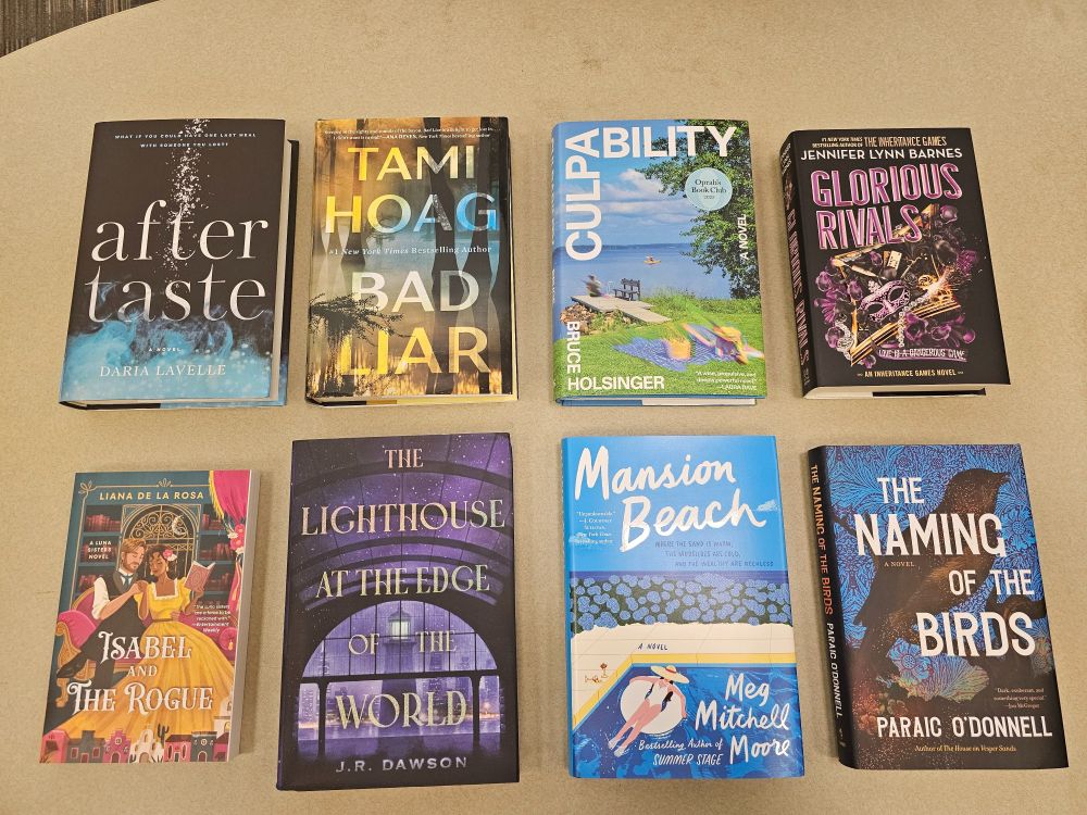 Eight fiction books on a table, from upper left: Aftertaste by Daria Lavelle,  Bad Liar by Tami Hoag, Culpability by Bruce Holsinger, Glorious Rivals by Jennifer Lynn Barnes, Isabel and the Rogue by Liana de la Rosa, The Lighthouse at the Edge of the World by J.R. Dawson, Mansion Beach by Meg Mitchell Moore, and The Naming of the Birds by Paraic O'Donnell.