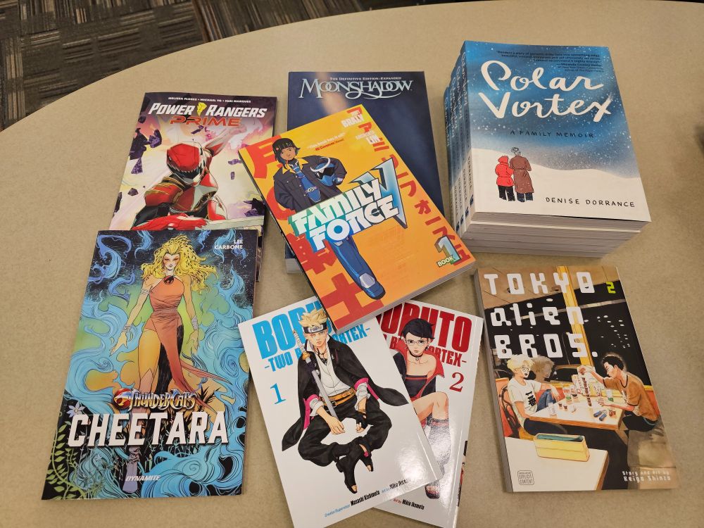 Eight graphic novels on a table from top left: Power Rangers Prime volume one, Moonshadow, Polar Vortex (5 copies for upcoming bookclub), in the center is Family Force V volume 1, bottom row from left starts with Thundercats Cheetara, Boruto Two Blue Vortex volumes one and two, and Tokyo Alien Bros. volume two.