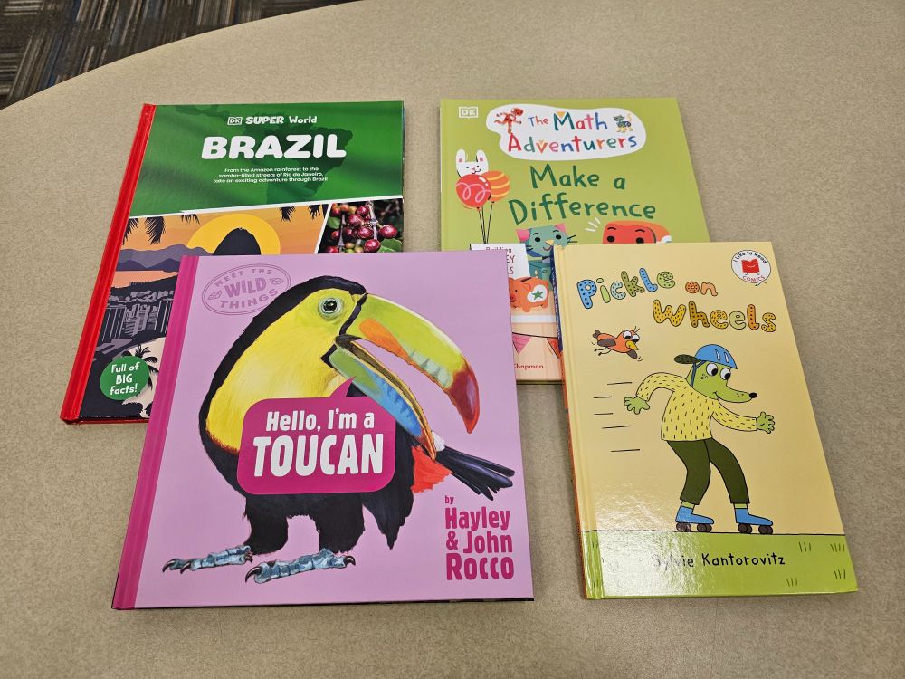 Four children's books on a table, from top left: DK Super World Brazil, The Math Adventurers Make a Difference by Sital Gorasia Chapman with Susanna Rumiz, Hello I'm a Toucan by Hayley and John Rocco, and Pickle on Wheels by Sylvie Kantorowitz.