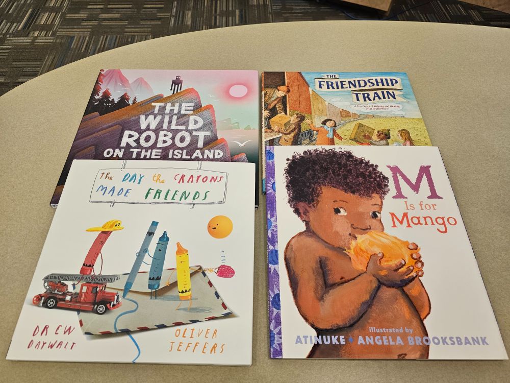 Four children's books on a table, from top left: The Wild Robot on the Island by Peter Brown, The Friendship Train by Debbie Levy with Boris Kulikov, The Day the Crayons Made Friends by Drew Daywalt with Oliver Jeffers, and M is for Mango by Atinuke with Angela Brooksbank.