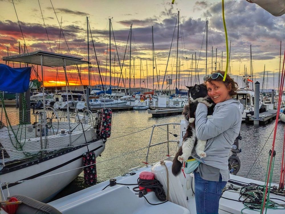 A woman (me) stands on the deck of a sailboat in a marina.  It is sunset, and the sky is a mix of orange, pink, and purple hues.  Many other sailboats are moored in the marina. I'm wearing a light gray jacket, blue jeans and aqua sunglasses on my head and holding Pseudo, a black and white cat close to my chest. A variety of nautical equipment, including ropes and other gear, is visible on the deck.  The water in the marina is a calm dark gray/blue. The image has a warm, inviting atmosphere although it's a chilly summer night. 