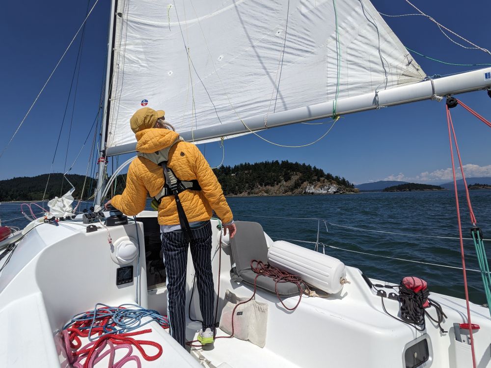 a man in the cockpit of a sailboat looking up at the mainsail. he has a yellow coat and a yellow hat on. 