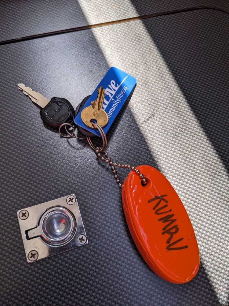 A close-up view of a set of keys, fobs, and a keychain on a dark gray textured surface. The keychain is orange colored, oval-shaped, and floating and has the word "KUMRU" written on it in black lettering.
