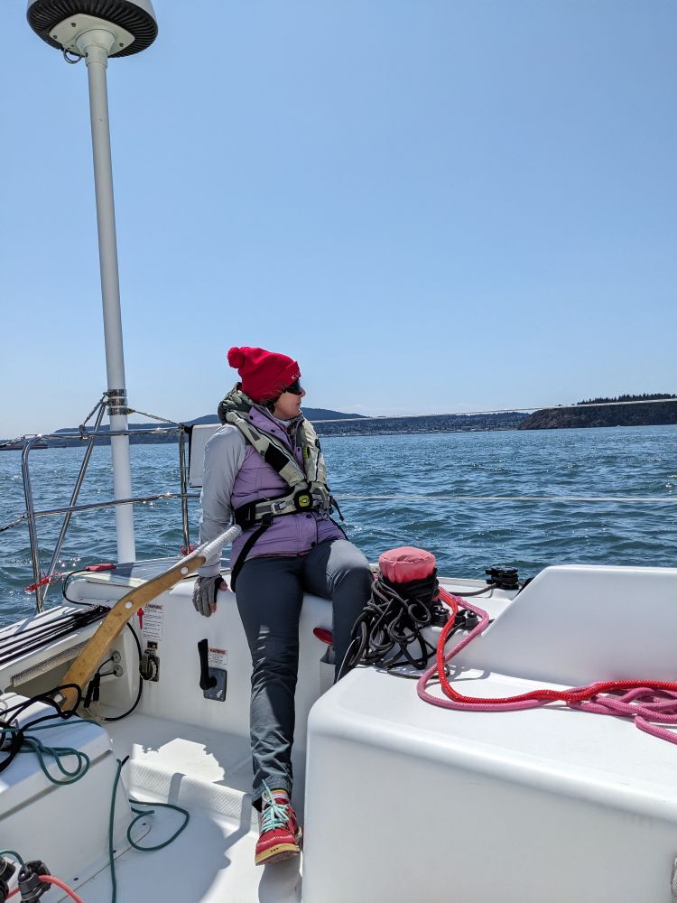 me sitting at the helm wearing a grey jacket and a lavender vest on top as well as a red hat. It's a gorgeous sunny day. 