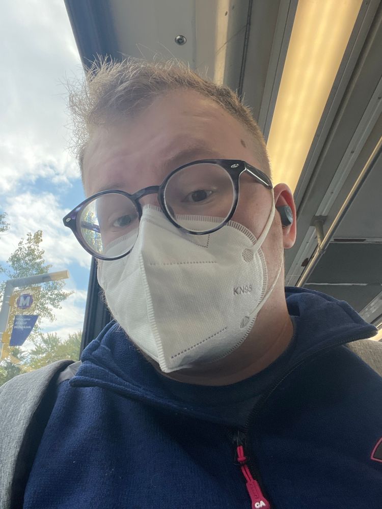 elliot wearing a mask on public transit. he has blue circular glasses on. he’s wearing a navy blue quarter zip. 