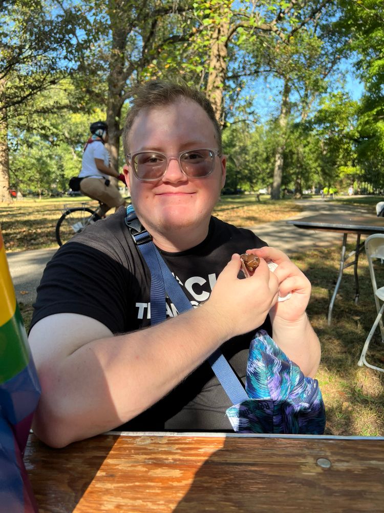 elliot holding an otter fidget. he’s got short brown hair and his facial hair is glowing the sun. he’s wearing clear glasses, a shirt that says fuck transphobia, and a blue and purple kavu shoulder bag. 