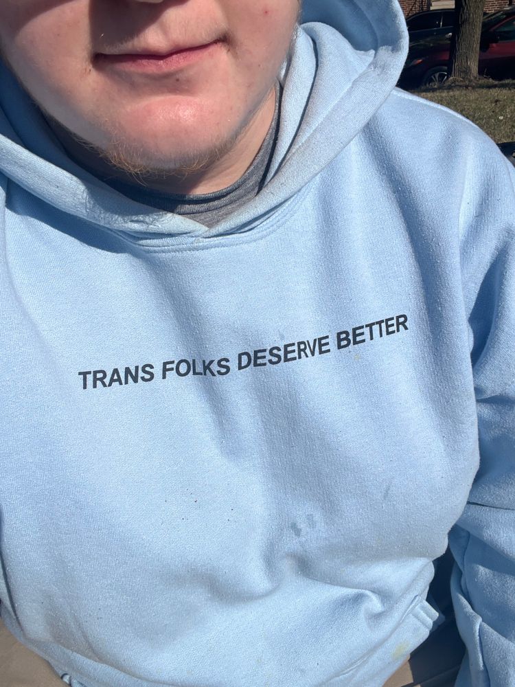 elliot wearing a baby blue hoodie that says trans folks deserve better. 