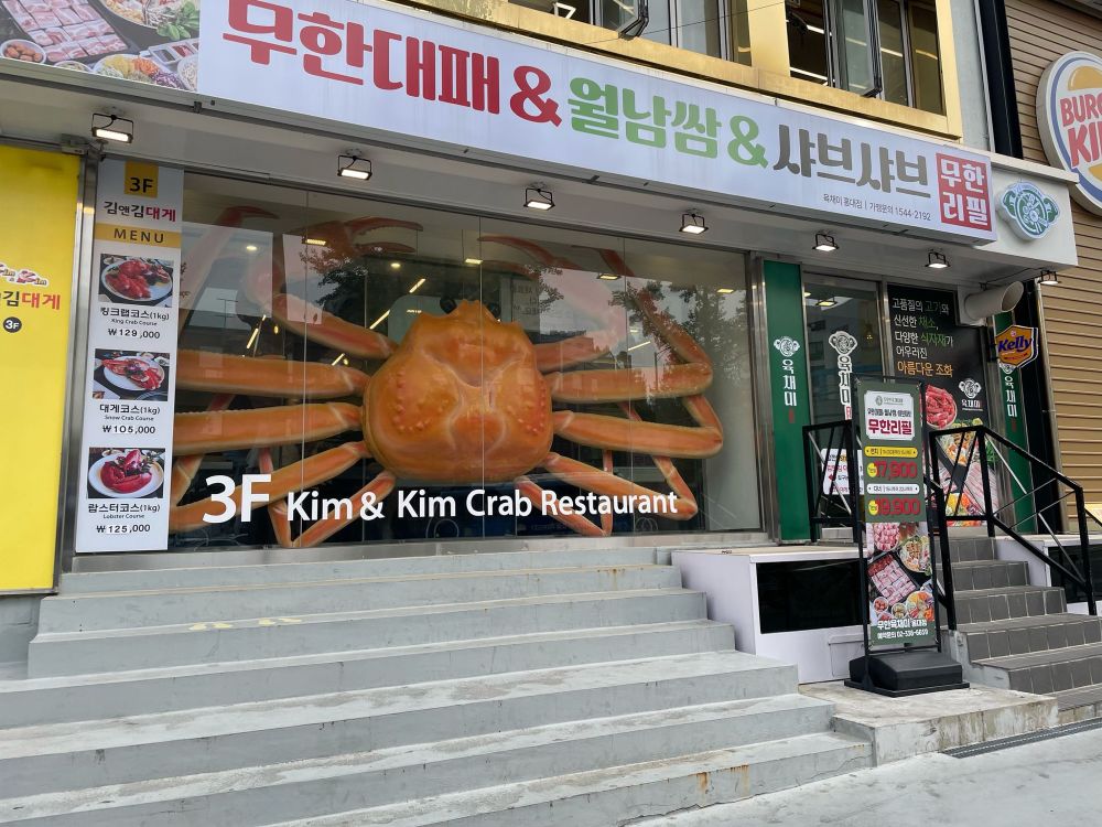 The widow of Kim & Kim Crab Restaurant in Seoul. There are concrete steps leading up to a plate glass window, behind which is a large crab.
“Large” is understating it. This crab could blot out the sun.
