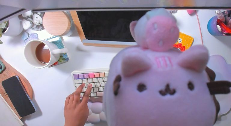 a picture of raynna's desk from a bird's eye view with a strawberry creme roll pusheen plushie close in frame on the right side of the screen.

the desk has their phone, a cup of coffee, various trinkets and crafts, and her keyboard set up on top.