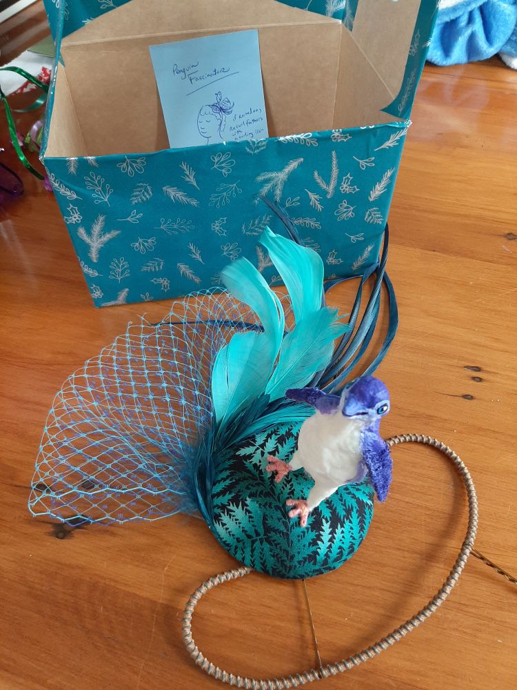 An incredible penguin fascinator! Blue netting and flowers with a blue and white penguin sitting on blue and black fabric. A cute drawing of a girl wearing a fascinator is on the box beside.