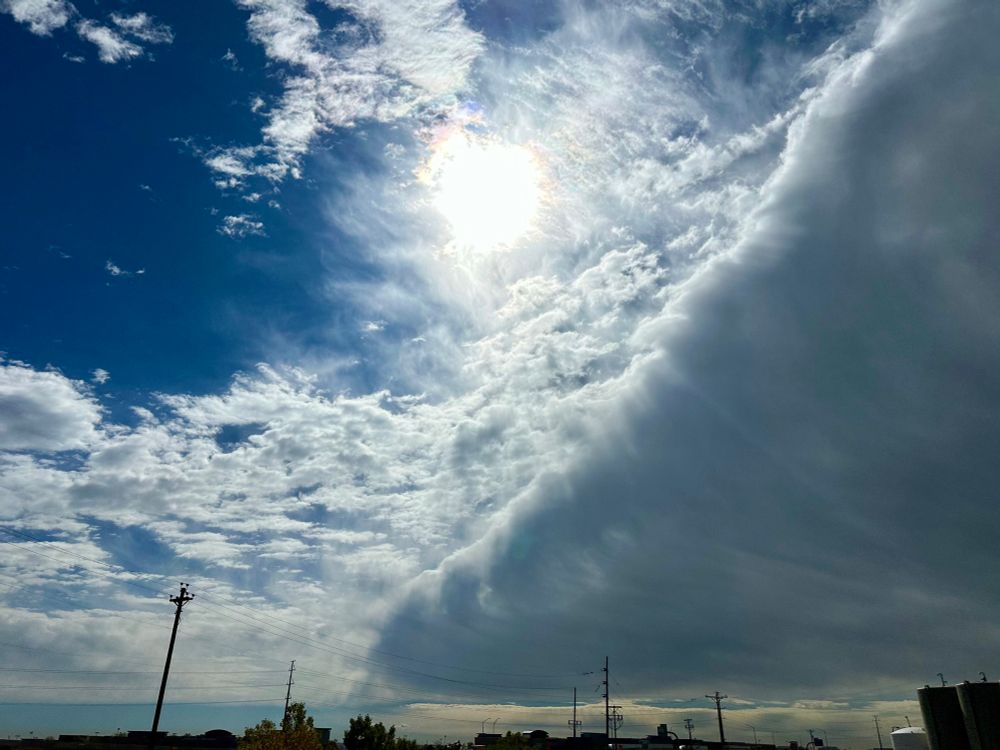 Photo of a massive cloud spread diagonally across the sky and the sun shining brightly behind a scattering of more clouds.