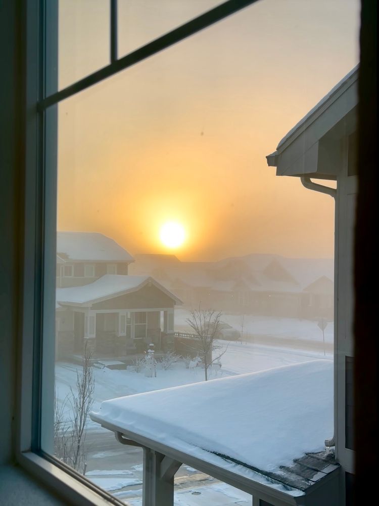 Photo through a window, of a foggy and snowy neighborhood with the sun rising as a blurry gradient of light through the fog.