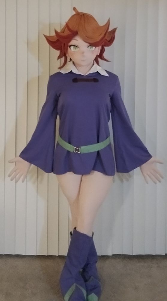 A kigurumi cosplayer dressed as Amanda O'Neill from Little Witch Academia, wearing the flight inform. She is standing in front of a white backdrop, facing the camera with legs crossed and arms out at her sides.