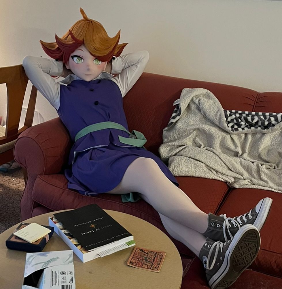 A kigurumi cosplayer dressed as Amanda O'Neill from Little Witch Academia, wearing her school uniform and converse shoes. She is reclining on a couch with her hands behind her head, legs crossed and resting on a footrest, looking at the camera.