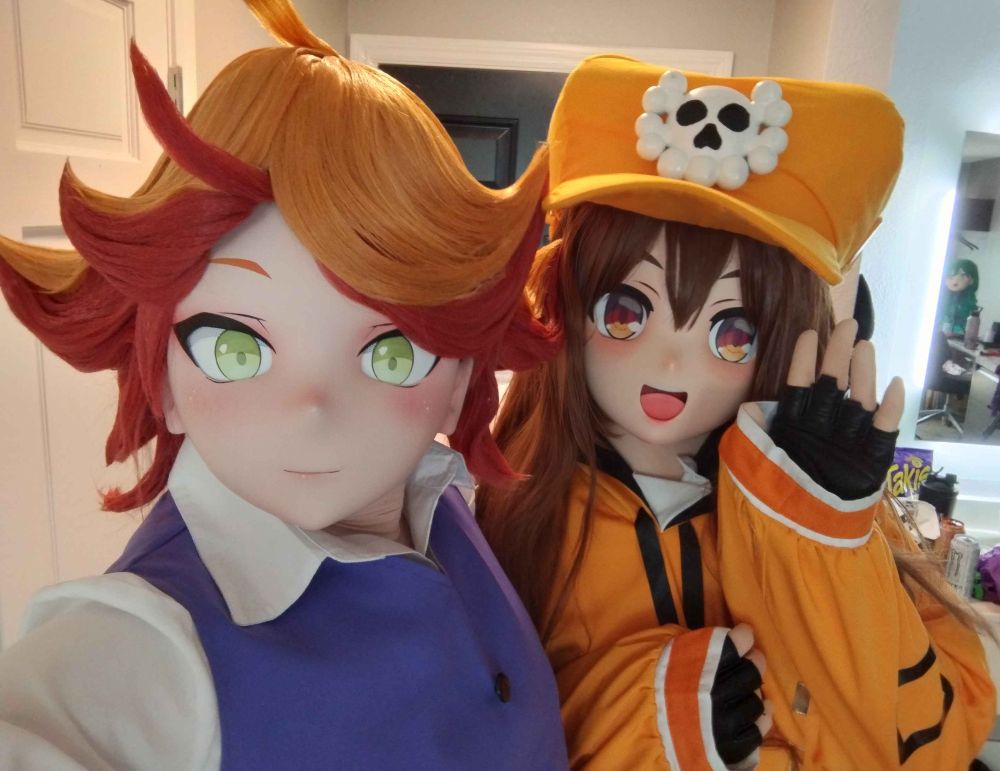 Two kigurumi cosplayers, one dressed as Amanda O'Neill from Little Witch Academia, the other dressed as May from Guilty Gear, taking a selfie.