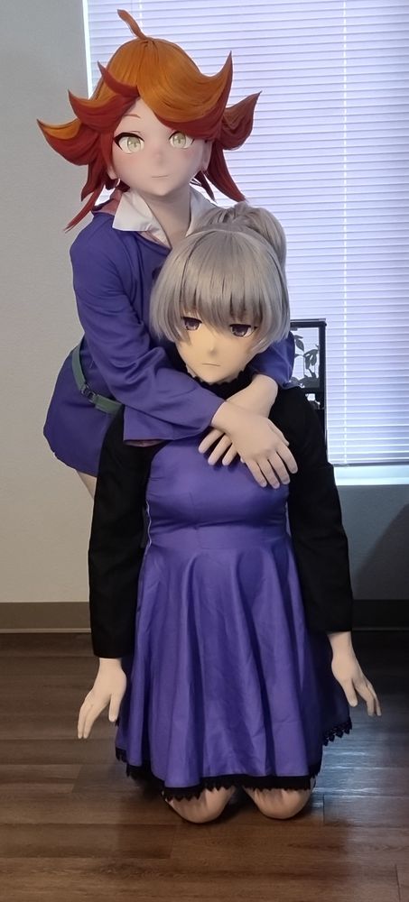 The same two kigurumi cosplayers. Yin is now kneeling on the floor, and Amanda is standing behind her, leaning forward with her arms loosely around Yin's shoulders.