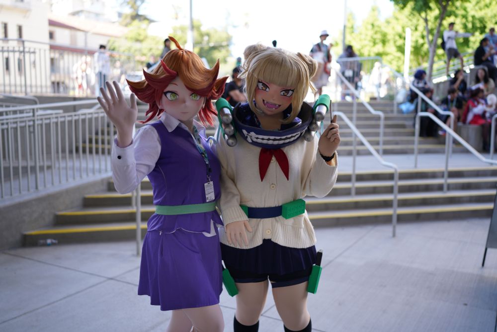 Two kigurumi cosplayers posing for  a photo outside Fanime. The one on the left is dressed as Amanda O'Neill from Little Witch Academia, wearing her school uniform. The one on the right is dressed as Himiko Toga from My Hero Academia, wearing her middle school sweater, big jaws bloodsucking thing that I don't know what it's called around her neck, and holding a knife in one hand.