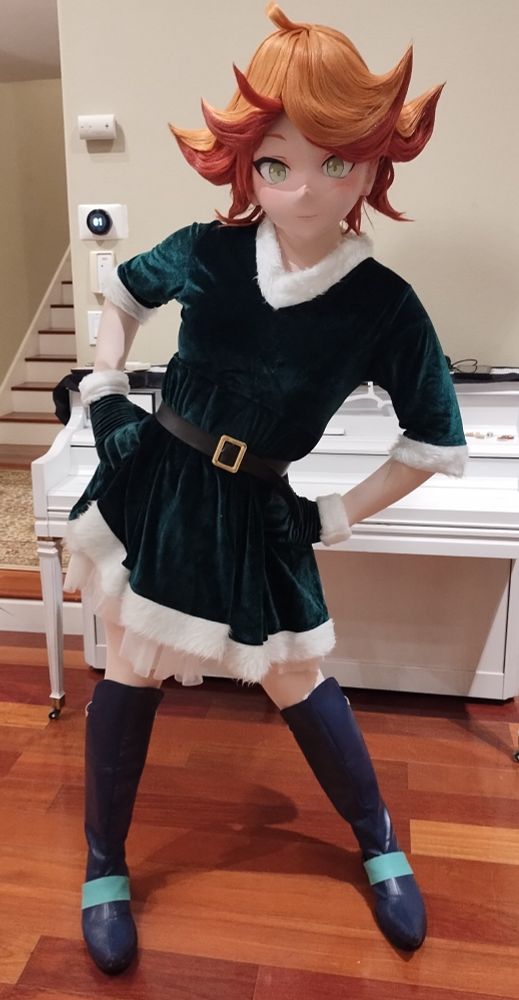 A kigurumi cosplayer dressed as Amanda O'Neill from Little Witch Academia, wearing a green Christmas-turned dress and blue boots. She is standing in front of a piano and has her hands on her hips.