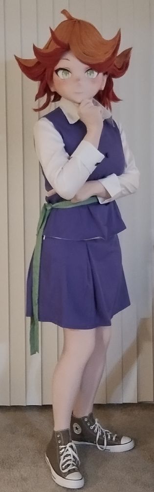 A kigurumi cosplayer dressed as Amanda O'Neill from Little Witch Academia, wearing her school uniform. She has one hand on her chin and is looking at the camera.