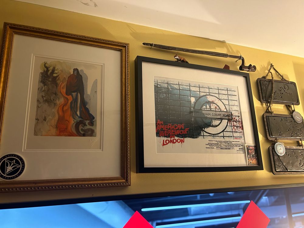 An image of two framed pieces of art on a yellow painted wall.  One is a Dalí woodblock watercolor print based on Dante’s Inferno.  One is a fanart promo poster for American Werewolf in London by Heather Vaughan.  Above the Vaughan print is a mounted civil war era bayonet. 