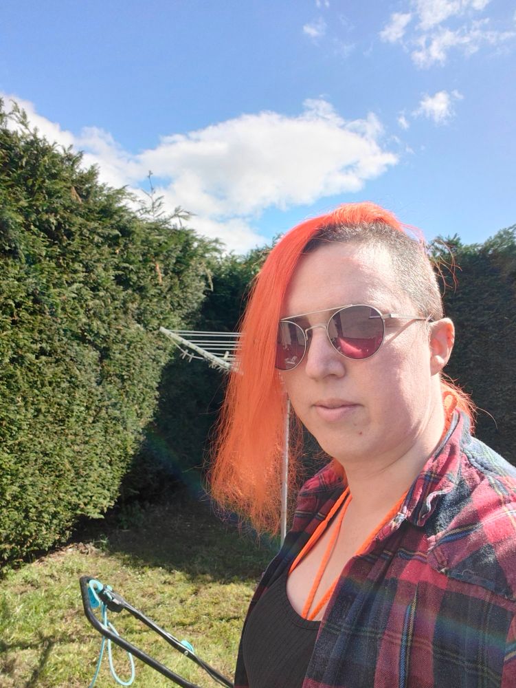Woman with orange hair in sunglasses, with hedges behind