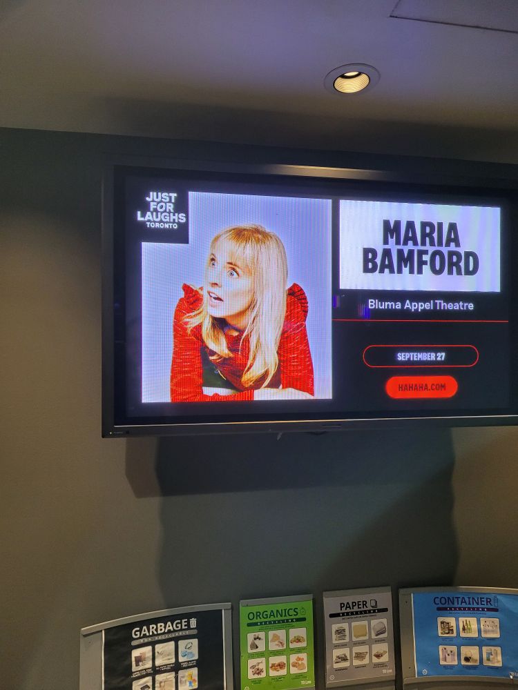 A tv screen with a picture of Maria Bamford looking to the side with a surprised expression. She has blonde hair and a red blouse. The screen also says Just for laughs Toronto. Maria Bamford, Bluma Appel Theatre, September 27, hahaha.com. 