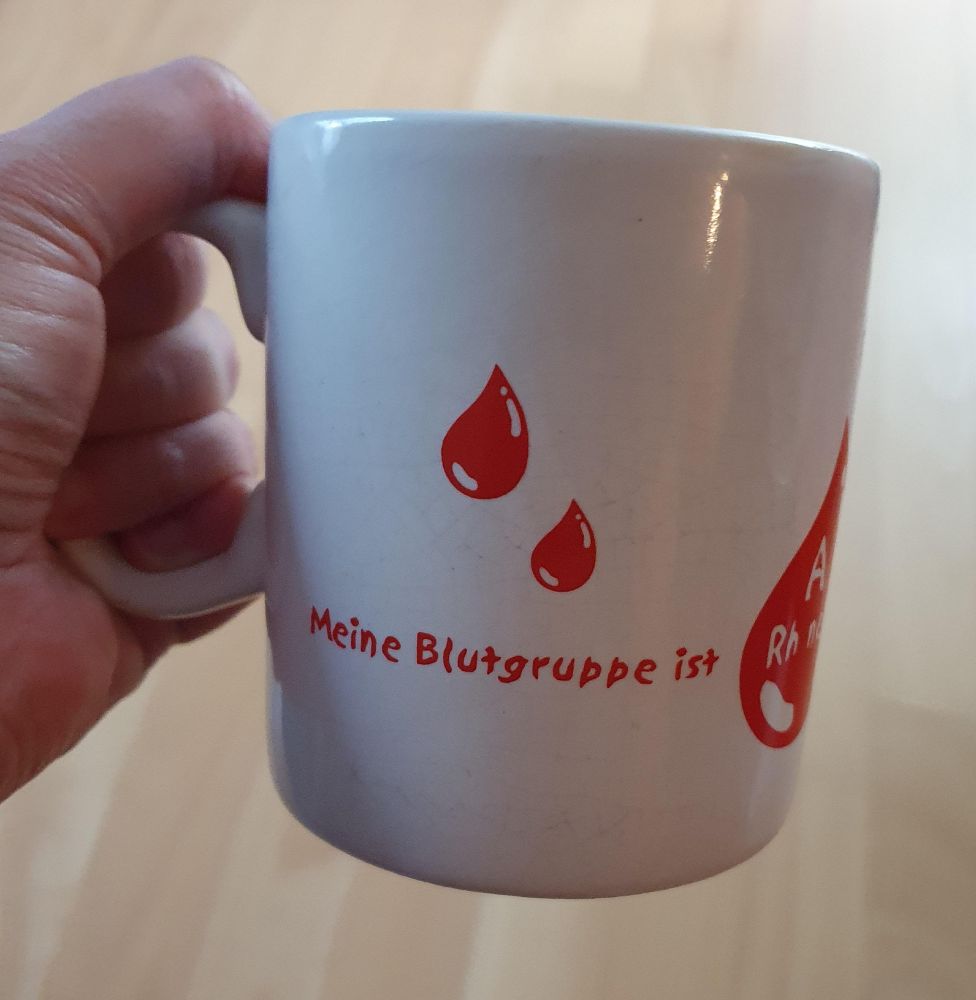 A hand holding a white mug. There's red blood drops on the mug and writing in German saying "Meine Blutgruppe ist", which means " my blood type is". Then there's a bigger red drop in which the blood type is written but you can't really see that part of the mug.