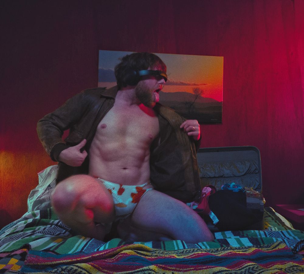Guy wearing leather jacket, cowprint briefs, with band over eyes, showing torso in a pose with stuck-out tongue