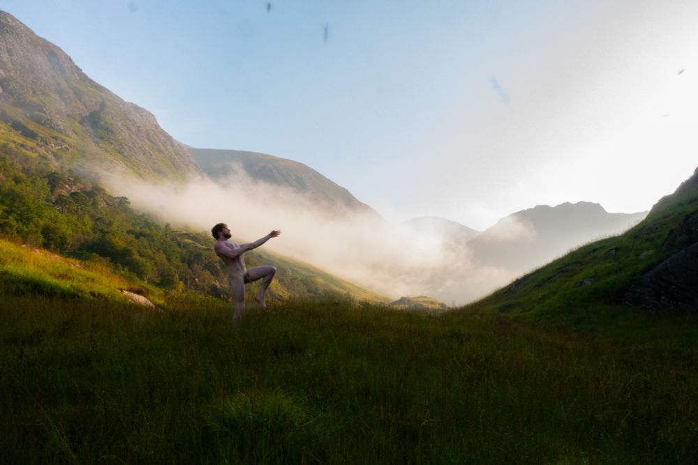 Sunrise strikes mist that lies on the green hills, while a nude man reaches out in the midground