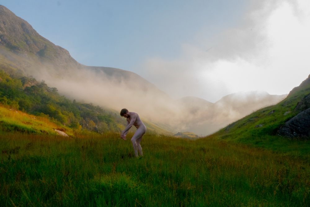 Sunrise strikes mist that lies on the green hills, while a nude man collapses inward in the midground