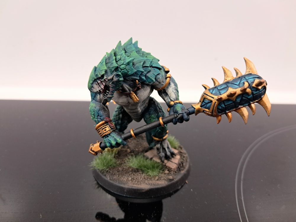 Photograph of a fully painted kroxigor. They're striding through jungle vegetation and are themselves a sea green/teal with pale bellies. Their metallic ornamentation and weapon details are a vibrant golden material, and their celestite weapons are a translucent turquoise crystal.
Their face is turned to display their nasty facial scarring, presumably caused by some huge, three-clawed beast.