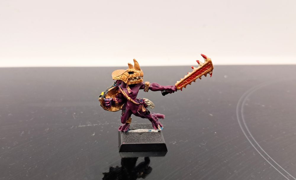 Photograph depicting a classic metal skink priest with a Macuahuitl.
Thus version was painted by a ten year old, with dark purple skin, black scales and with a golden mask and weapon. The weapon is, predictably, covered in red blood.