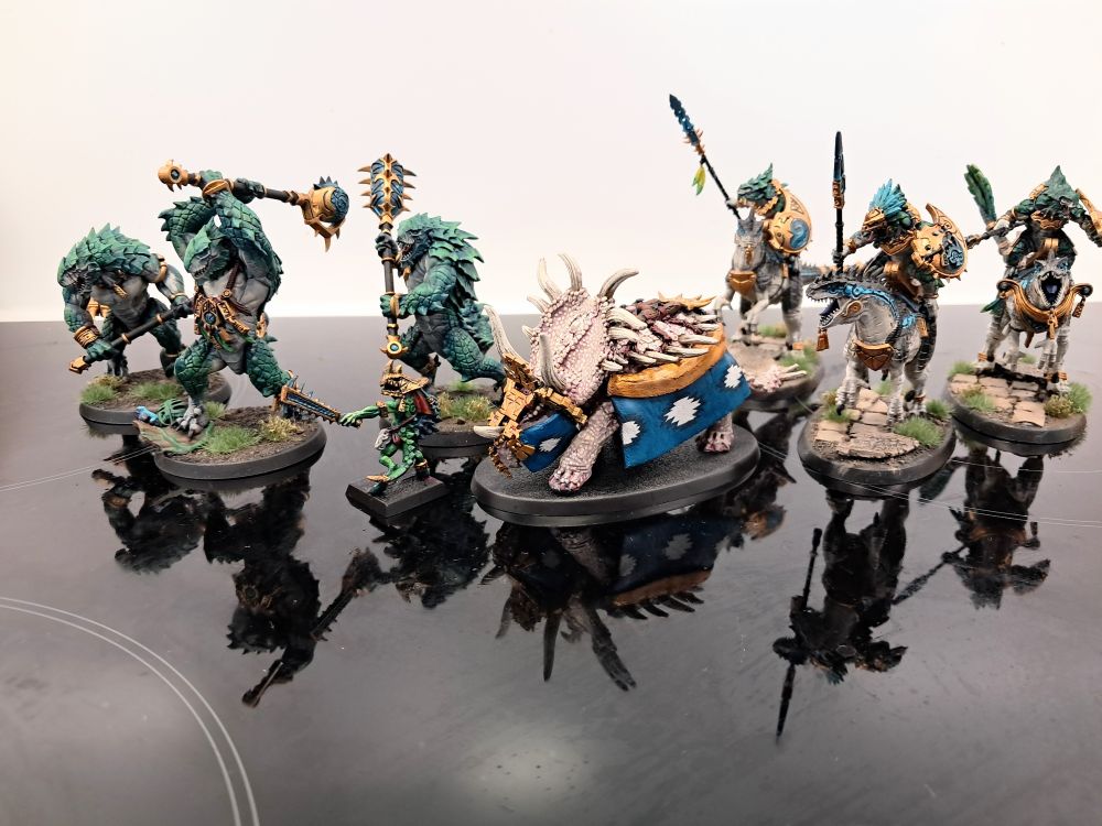 Photograph depicting a classic metal stegadon, next to a classic metal skink priest, a unit of kroxigor and a unit of aggradon riders from Warhammer. The classic models are from the old warhammer fantasy range, the others are from Age of Sigmar.
The fully painted riders, kroxigor and skink are teal/green, with more vibrant green on the smaller, softer skink and more blue-green on the larger, heavily armoured kroxigor. The kitbashed skirts of the stegadon are teal, to match the scheme of the other seraphim, on account of being otherwise albino